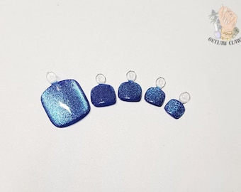 May include: A set of five square-shaped press-on toenails in a vibrant royal blue color with a dense glitter finish. Each nail features a small, clear plastic tab at the base for application. The logo Outlaw Claws is visible in the top right corner.
