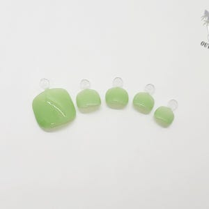 May include: A set of five light green press-on toenails, each with a rounded square shape and a small loop at the top. The nails are arranged in a row, decreasing in size from left to right. The background is plain white.