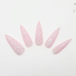 May include: Five light pink, stiletto-shaped press-on nails with white floral and heart designs. The nails are arranged in a fan shape against a white background. The Outlaw Claws logo is in the upper right corner.