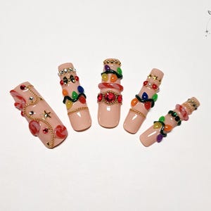 May include: Five artificial nails with a nude base color, each adorned with intricate designs. Decorations include colorful gems, gold chains, and miniature Christmas lights. The nails are arranged on a white surface.