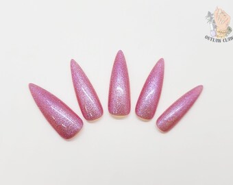 Puede incluir: Cinco uñas postizas rosas con purpurina dispuestas en forma de abanico. Las uñas son largas y puntiagudas, con un acabado brillante e iridiscente. El logotipo de Outlaw Claws está en la esquina superior derecha.