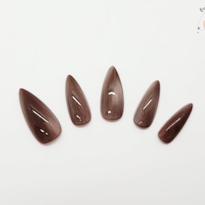 May include: Five almond-shaped, glossy, brown press-on nails arranged on a white surface. The nails have a subtle cat-eye effect, with a lighter streak down the center. The Outlaw Claws logo is in the upper right corner.