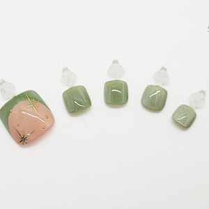May include: A set of five artificial toenails. One nail features a pink and green design with gold star accents. The other four nails are a solid green color. Each nail is attached to a clear plastic holder.