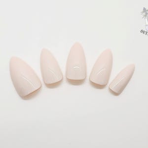 May include: Five almond-shaped, pale pink press-on nails arranged on a white surface. The nails have a glossy finish and are evenly spaced. The Outlaw Claws logo is in the upper right corner.