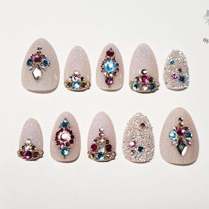May include: A set of ten almond-shaped, glittery light pink press-on nails. Each nail is adorned with colorful rhinestones in various designs, including clusters and geometric patterns. The Outlaw Claws logo is visible in the upper right corner.