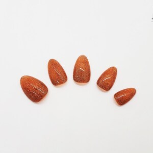 May include: Set of six almond-shaped, press-on nails in a warm, shimmering copper color. The nails are arranged on a white surface, showcasing their glossy finish and glittery texture. The Outlaw Claws logo is in the upper right corner.