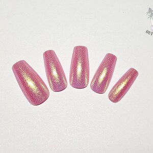 May include: Five press-on nails in a coffin shape, featuring a vibrant pink base color with a dense, shimmering gold glitter finish. The nails are arranged in a fan-like pattern, displaying a reflective, metallic-like texture across the surface.