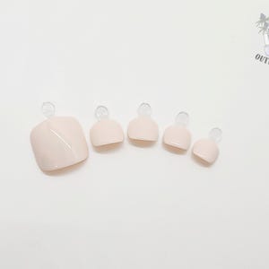 May include: A set of five light pink press-on toenails, varying in size, arranged on a white surface. Each nail has a rounded shape with a clear tab at the top. The Outlaw Claws logo is visible in the upper right corner.