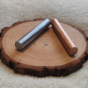 May include: Two metal rods, one silver and one copper, are placed on a wooden slice. The rods are approximately 15 cm long.