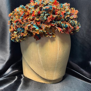 May include: A colorful, textured headpiece with a mix of red, teal, and gold fabric. The headpiece is displayed on a wooden head form against a black background. The fabric appears to be ruffled or layered, creating a voluminous effect.