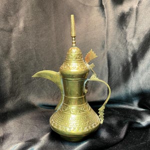May include: A vintage brass coffee pot with intricate engraved patterns. The pot features a long spout, a curved handle, and a decorative finial on the lid. The brass has a warm, aged patina, suggesting its antique nature. The pot is likely used for serving coffee or tea.