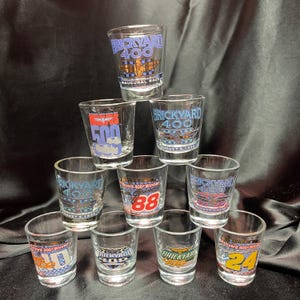 May include: A collection of clear glass shot glasses. Each glass features a different design related to the Brickyard 400 race, including logos, numbers, and the text "Inaugural Race". The glasses are stacked in a pyramid shape.