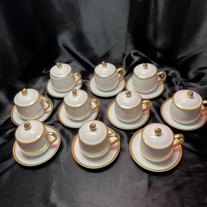 May include: A set of ten white porcelain espresso cups with matching saucers. Each cup has a gold handle, gold trim, and a small lid with a gold knob. The saucers also have gold trim. The cups are arranged on a dark background.