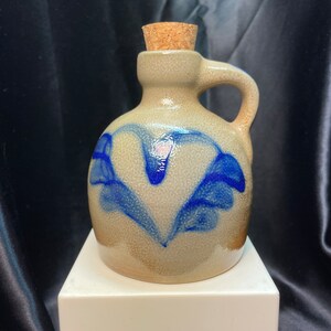 May include: A small, gray stoneware jug with a blue heart design. The jug has a cork stopper and a handle.
