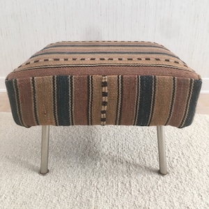 May include: A rectangular ottoman with a woven fabric top featuring vertical stripes in brown, tan, and navy blue. The ottoman has four silver metal legs and is placed on a white rug.