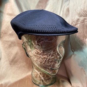 May include: A navy blue flat cap with a textured knit design. The cap is displayed on a clear glass mannequin head. The cap has a short brim and a rounded crown. The background is a light brown fabric.