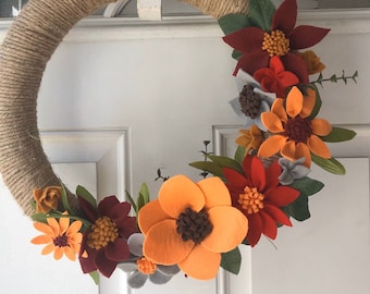 May include: A fall-themed wreath made with burlap rope and felt flowers in shades of orange, red, and burgundy. The wreath is decorated with green leaves and small brown felt flowers.