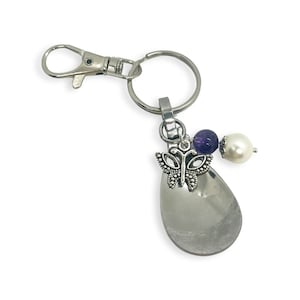 May include: A silver key chain with a clear teardrop-shaped charm, a silver butterfly charm, a purple amethyst bead, and a white pearl bead.