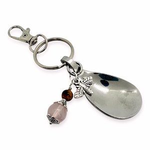 Może przedstawiać: Brelok do kluczy w kształcie srebrnej łyżeczki z zawieszką z różowo-brązową koralikiem, srebrnym zawieszką w kształcie ważki i srebrnym rozdzielonym pierścieniem.