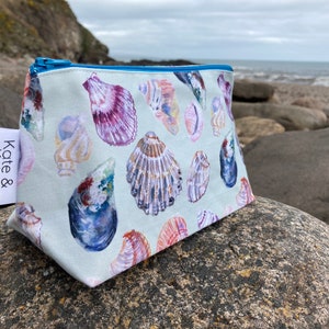 May include: A light blue zippered pouch with a white background and a pattern of colourful seashells. The pouch is sitting on a rock with a blurry background of a beach and ocean. The text "Kate & Kelpie" is printed on a white label attached to the pouch.