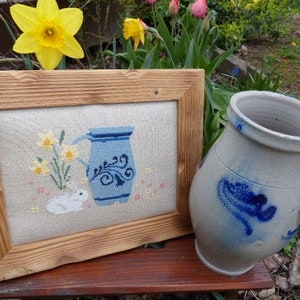 Può includere: Un ricamo a punto croce incorniciato che rappresenta una brocca blu con un coniglio bianco e fiori gialli. Una brocca in gres blu e bianco è in primo piano.