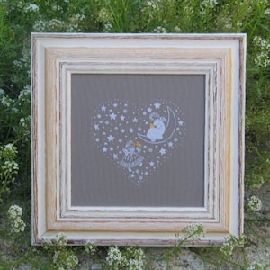 May include: A framed cross-stitch artwork featuring a heart-shaped design filled with stars. The artwork depicts a figure on a crescent moon and another figure below. The frame is a distressed white and tan color, set against a backdrop of greenery.