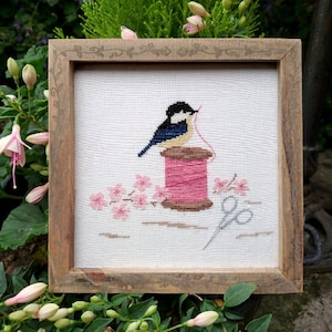 May include: A cross-stitch embroidery of a bluebird perched on a spool of pink thread. The bird is holding a red thread in its beak. There are pink flowers and a pair of silver scissors in the background.