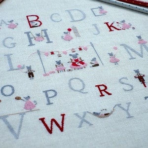 May include: A white fabric with a cross-stitch alphabet design featuring gray and red letters and small mouse illustrations. The letters are arranged in alphabetical order and each letter is paired with a mouse doing a different activity.