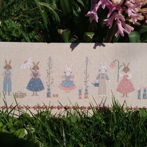 May include: Cross-stitch embroidery of a group of bunnies dressed in different outfits. The bunnies are holding flowers, watering cans, and a basket. The embroidery is on a beige linen fabric.