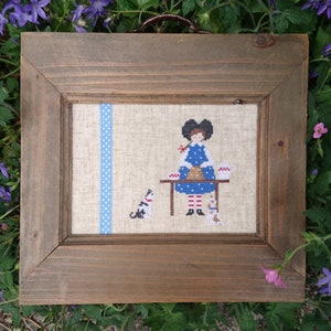 Peut inclure: Une broderie encadrée représentant une fille en robe bleue à pois blancs, debout derrière une table. Un chat et deux autres animaux sont également présents. Le cadre est en bois brun, et une bande bleue à pois longe le côté gauche.