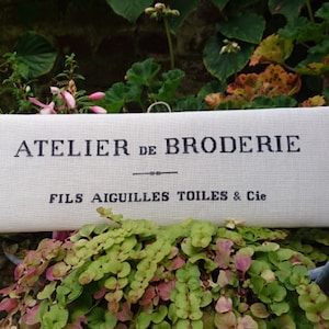 Puede incluir: Un letrero blanco con texto negro que dice "Atelier de Broderie Fils Aiguilles Toiles & Cie". El letrero está colgado de un gancho y está rodeado de plantas verdes.