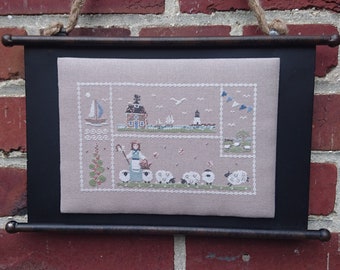 May include: A framed cross-stitch embroidery depicting a pastoral scene with a shepherdess, sheep, a house, a lighthouse, and a sailboat. The embroidery is on a beige linen fabric and is framed in a black wood frame.