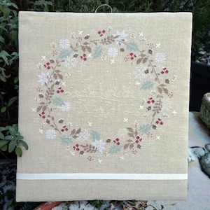 Peut inclure: Un tissu de lin beige avec un motif de couronne brodée en blanc. La couronne est composée de feuilles, de baies et de flocons de neige.
