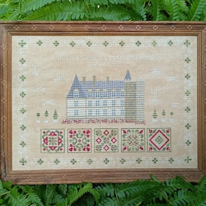 May include: A framed cross-stitch embroidery depicting a blue and white castle with a green roof. The castle is surrounded by a border of green trees and red, white, and blue flowers. The text "A Cottage of Delight" is stitched above the castle.