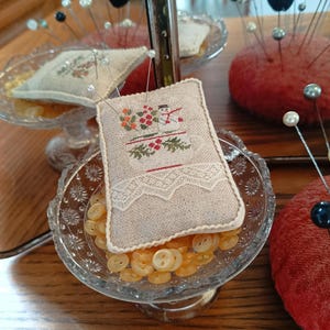 Puede incluir: Un pequeño cojín de alfileres rectangular beige con un diseño de punto de cruz de un muñeco de nieve y elementos florales, que descansa sobre un plato de vidrio lleno de botones amarillos. El plato se encuentra en un soporte escalonado.