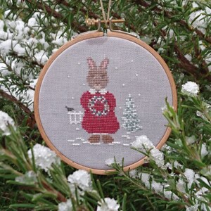 Puede incluir: Un adorno de aro bordado en blanco y gris con un conejito vestido de rojo que sostiene una corona. El conejito está de pie frente a un pequeño árbol verde. El fondo es gris claro con copos de nieve blancos.
