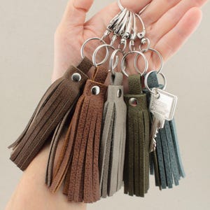 Könnte beinhalten: Eine Sammlung von fünf Leder-Quasten-Schlüsselanhängern in Dunkelbraun, Mittelbraun, Hellgrau, Olivgrün und Schieferblau. Jede Quaste hat einen silberfarbenen Druckknopf, einen Schlüsselring und einen Karabinerhaken. Ein silberner Schlüssel ist befestigt.