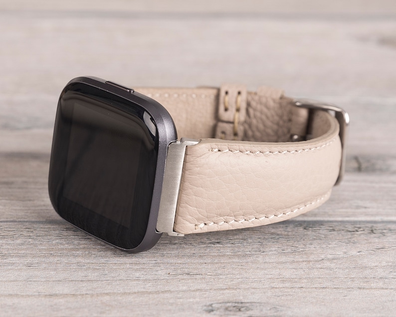 Puede incluir: Una correa de reloj de cuero beige con costuras blancas para un reloj inteligente. La correa se muestra en un reloj inteligente con una pantalla negra.