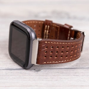 May include: A smartwatch with a dark gray case and a brown leather band. The band features a perforated design and is secured with a silver buckle. The watch is displayed on a light wood-grain surface.
