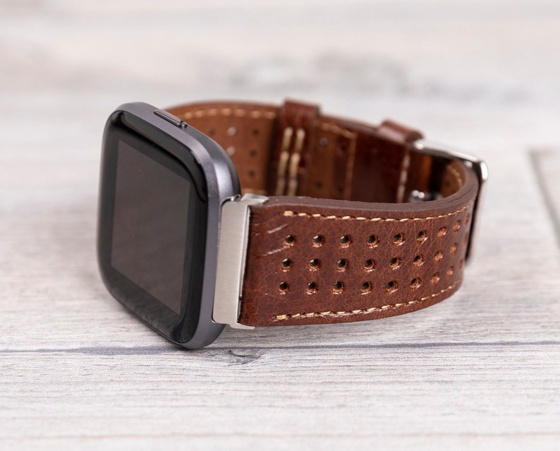 May include: A smartwatch with a dark gray case and a brown leather band. The band features a perforated design and is secured with a silver buckle. The watch is displayed on a light wood-grain surface.
