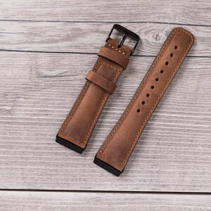 Puede incluir: Dos correas de reloj de cuero marr&oacute;n con herrajes met&aacute;licos negros. Las correas est&aacute;n dispuestas sobre una superficie de madera clara.