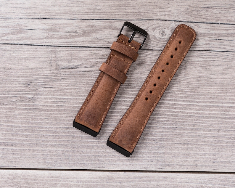 Puede incluir: Dos correas de reloj de cuero marr&oacute;n con herrajes met&aacute;licos negros. Las correas est&aacute;n dispuestas sobre una superficie de madera clara.