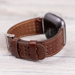 May include: Brown leather watch band with a perforated design and a silver buckle. The watch band is attached to a black smartwatch, showcasing a classic and sporty style. The band is made of genuine leather.