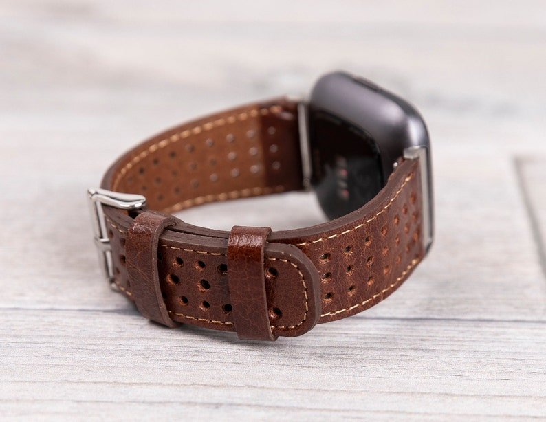 May include: Brown leather watch band with a perforated design and a silver buckle. The watch band is attached to a black smartwatch, showcasing a classic and sporty style. The band is made of genuine leather.