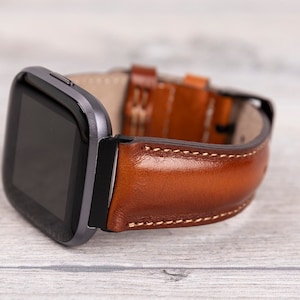 Könnte beinhalten: Ein braunes Lederarmband mit weißen Nähten. Das Armband ist an einer schwarzen Smartwatch mit quadratischem Zifferblatt befestigt.