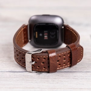 May include: Brown leather watch band with a perforated design and a silver buckle. The watch band is attached to a dark gray smartwatch, showcasing the back of the watch. The band has white stitching.