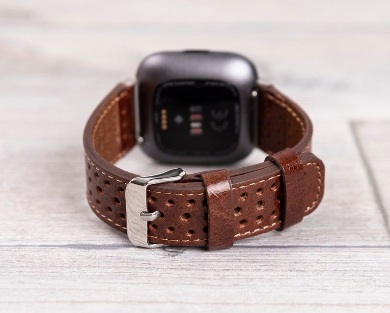 May include: Brown leather watch band with a perforated design and a silver buckle. The watch band is attached to a dark gray smartwatch, showcasing the back of the watch. The band has white stitching.