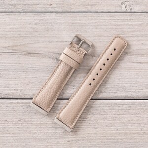 Puede incluir: Dos correas de reloj de cuero beige con hebillas plateadas. Las correas est&aacute;n dispuestas sobre una superficie de madera clara.