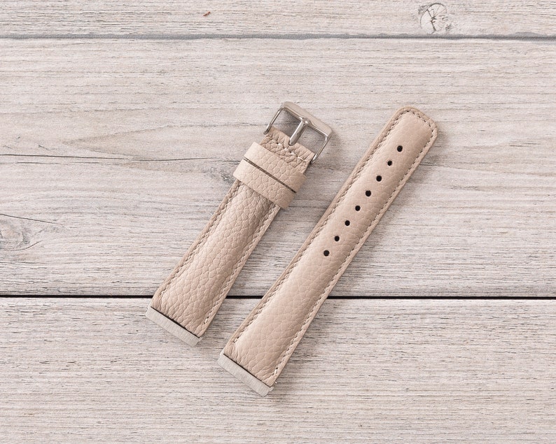 Puede incluir: Dos correas de reloj de cuero beige con hebillas plateadas. Las correas est&aacute;n dispuestas sobre una superficie de madera clara.
