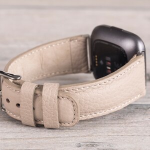 Puede incluir: Una correa de reloj de cuero beige con una hebilla plateada. La correa est&aacute; dise&ntilde;ada para un reloj inteligente con una cara cuadrada.